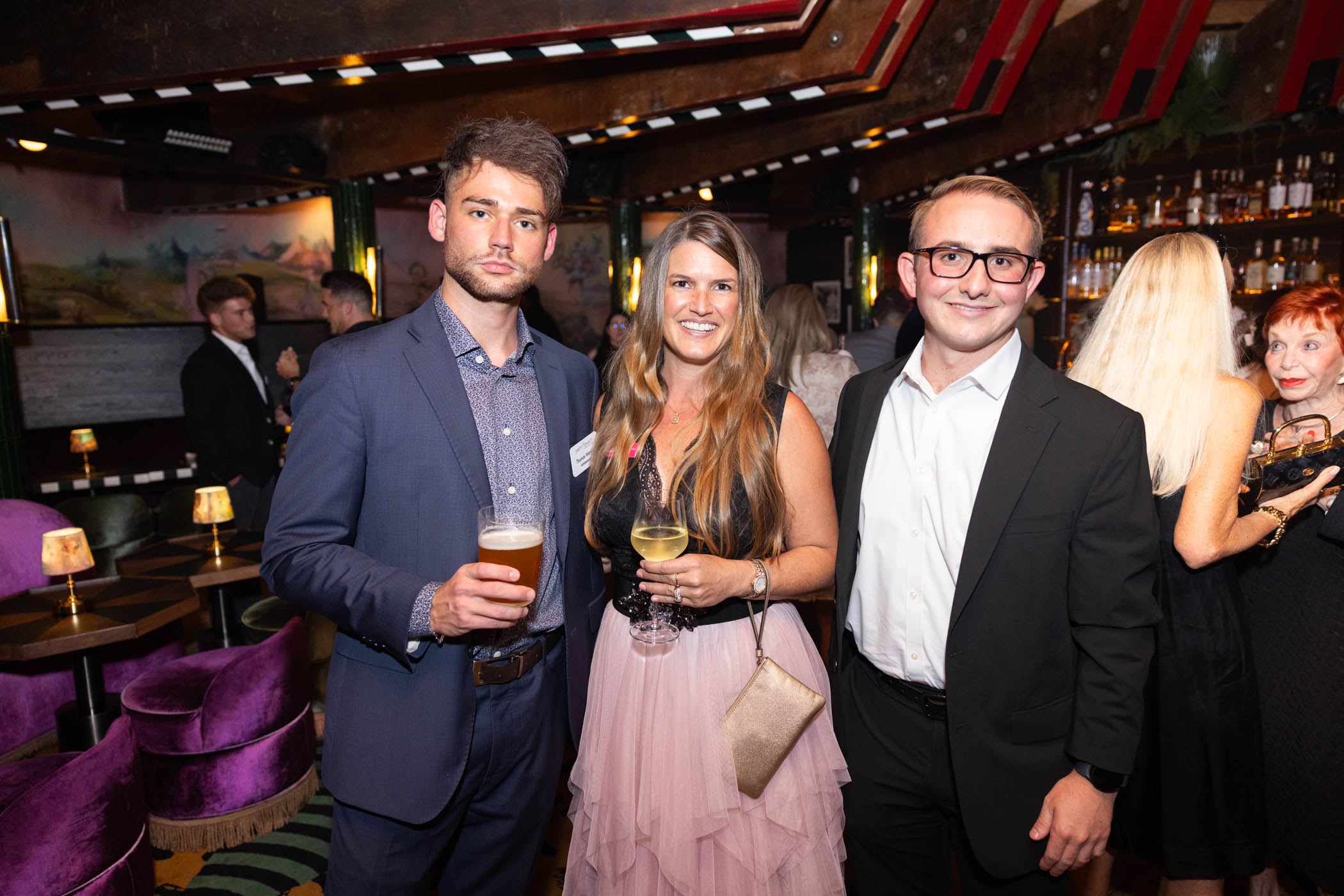 Three people dressed in formal attire pose and smile at a stylish indoor event, holding drinks. The room has velvet chairs, dim lighting, and others socializing in the background.