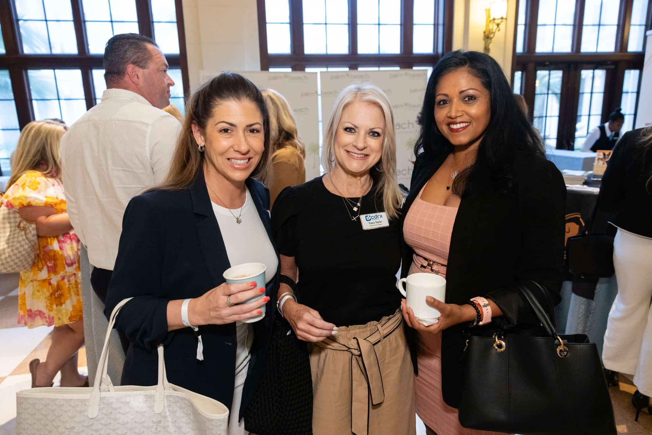 Three women stand together, smiling and holding coffee cups at an indoor event. Other people mingle in the background. The women are dressed in business attire and appear to be enjoying a professional gathering.