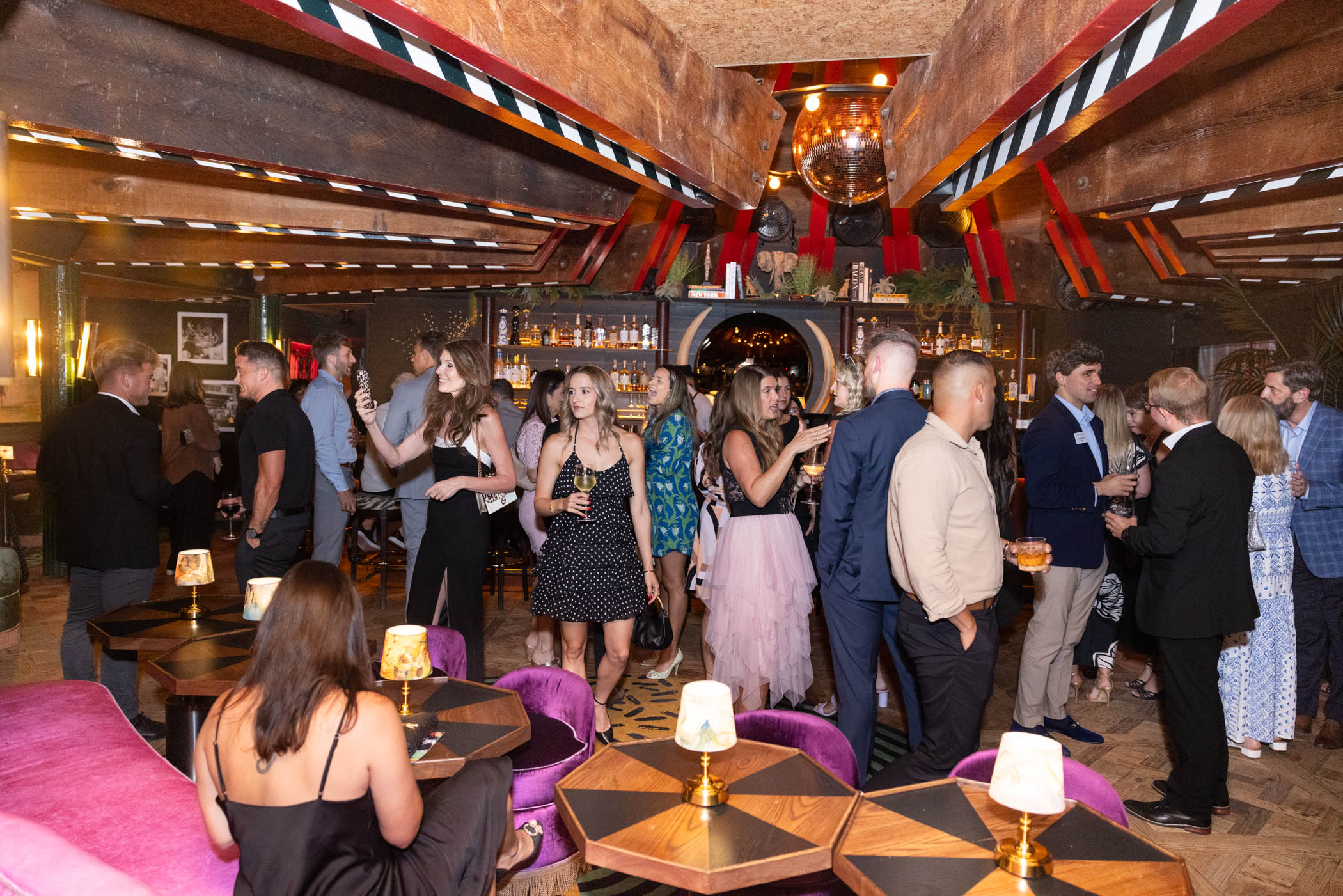 A group of well-dressed people mingle, chat, and hold drinks in a stylish, dimly lit bar with velvet chairs, small tables, hanging lights, and a mirror-backed bar in the background.