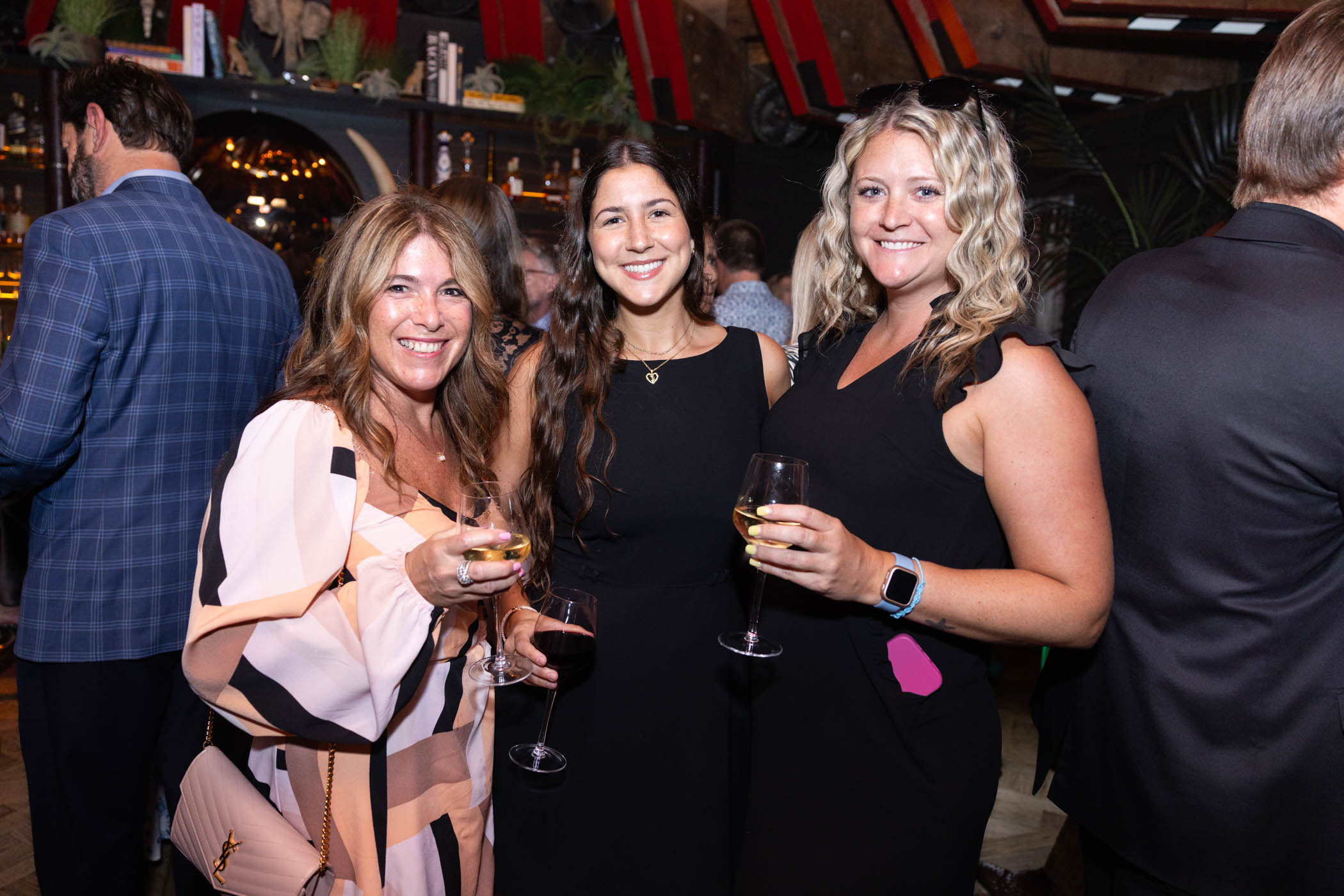 Three women dressed in black and patterned dresses smile while holding glasses of wine at an indoor social event; other guests mingle in the background near a bar.
