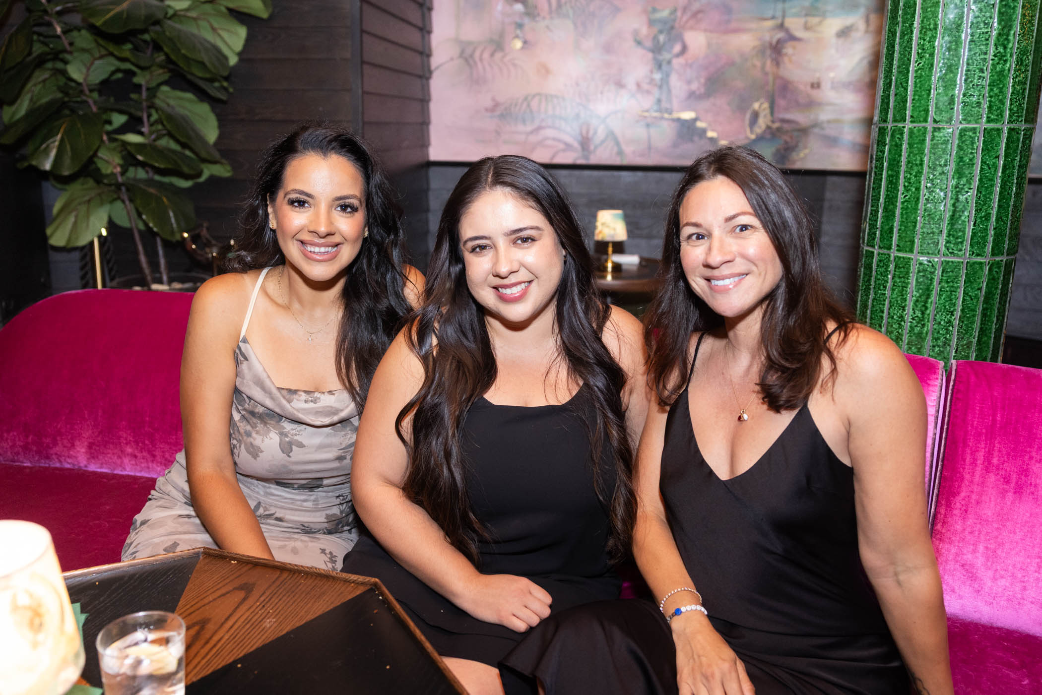 Three women are sitting on a bright pink couch in a stylish indoor setting, smiling at the camera. They are dressed in elegant evening dresses, and there are drinks and a small lamp on the table in front of them.