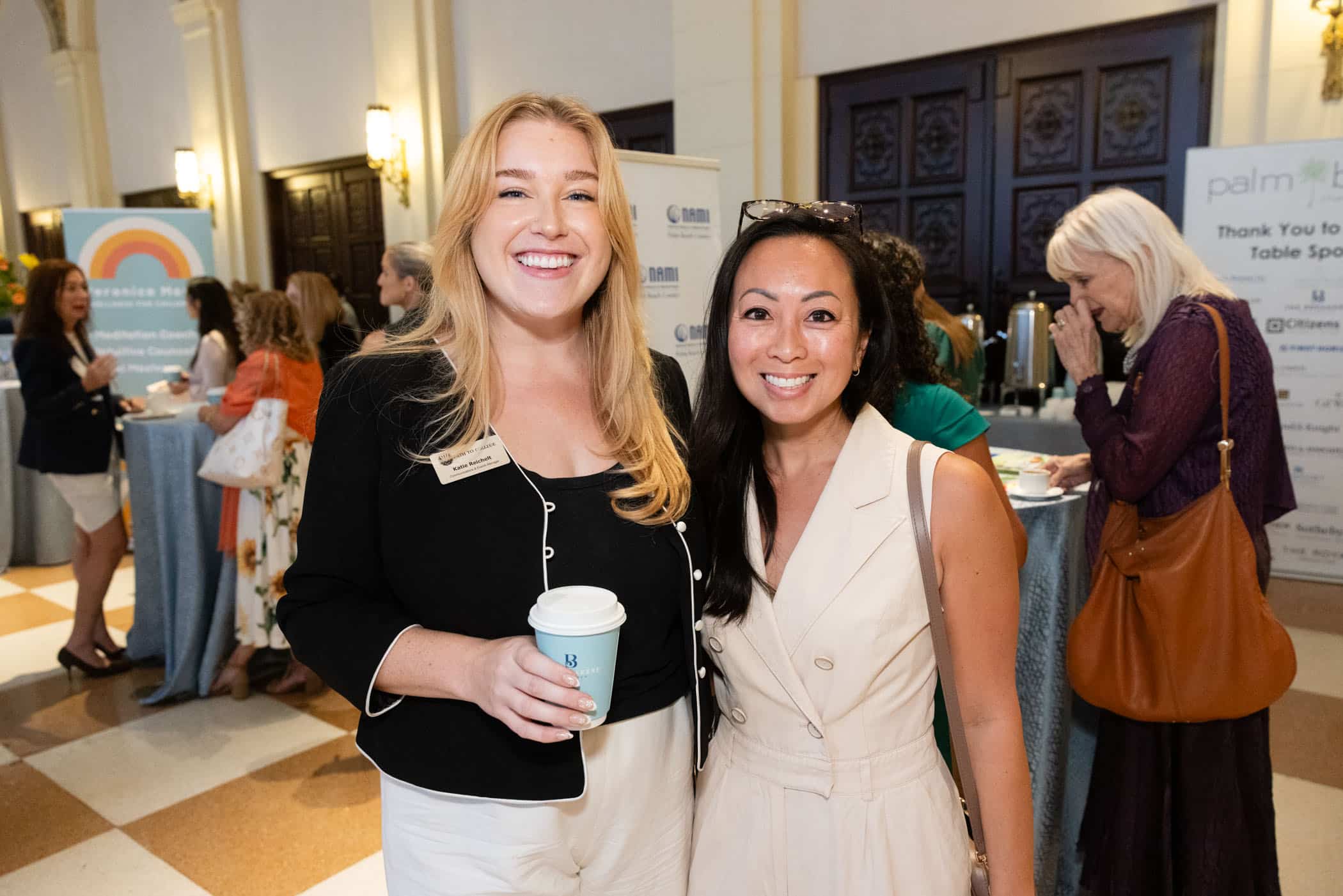Two women smiling and standing together at a networking event; one holds a coffee cup. People in the background are talking near round tables with brochures and banners displayed. The atmosphere is professional and welcoming.