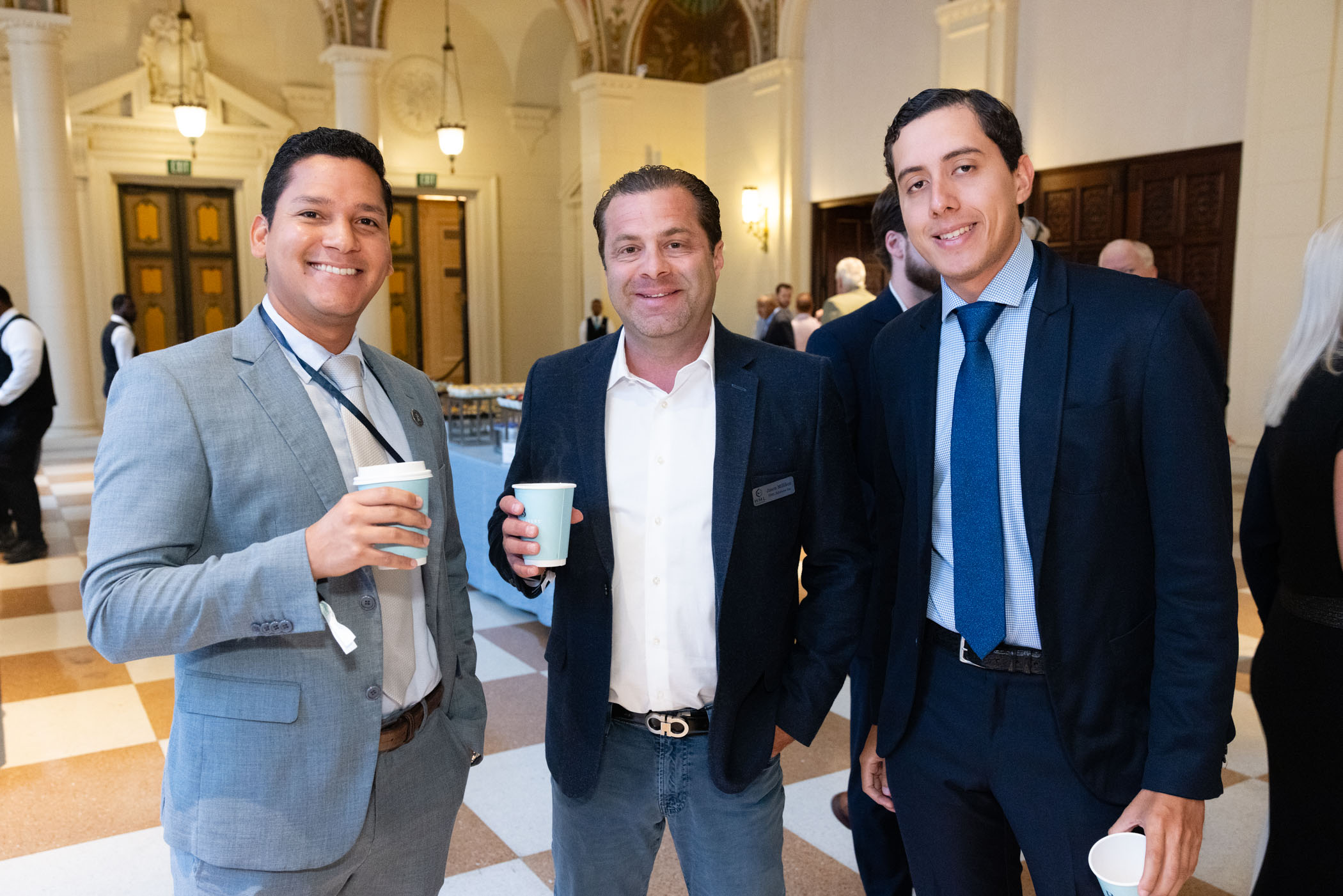 Three men in business attire stand together indoors, smiling at the camera and holding coffee cups. The background shows a large, elegant hall with checkered floors and people mingling.
