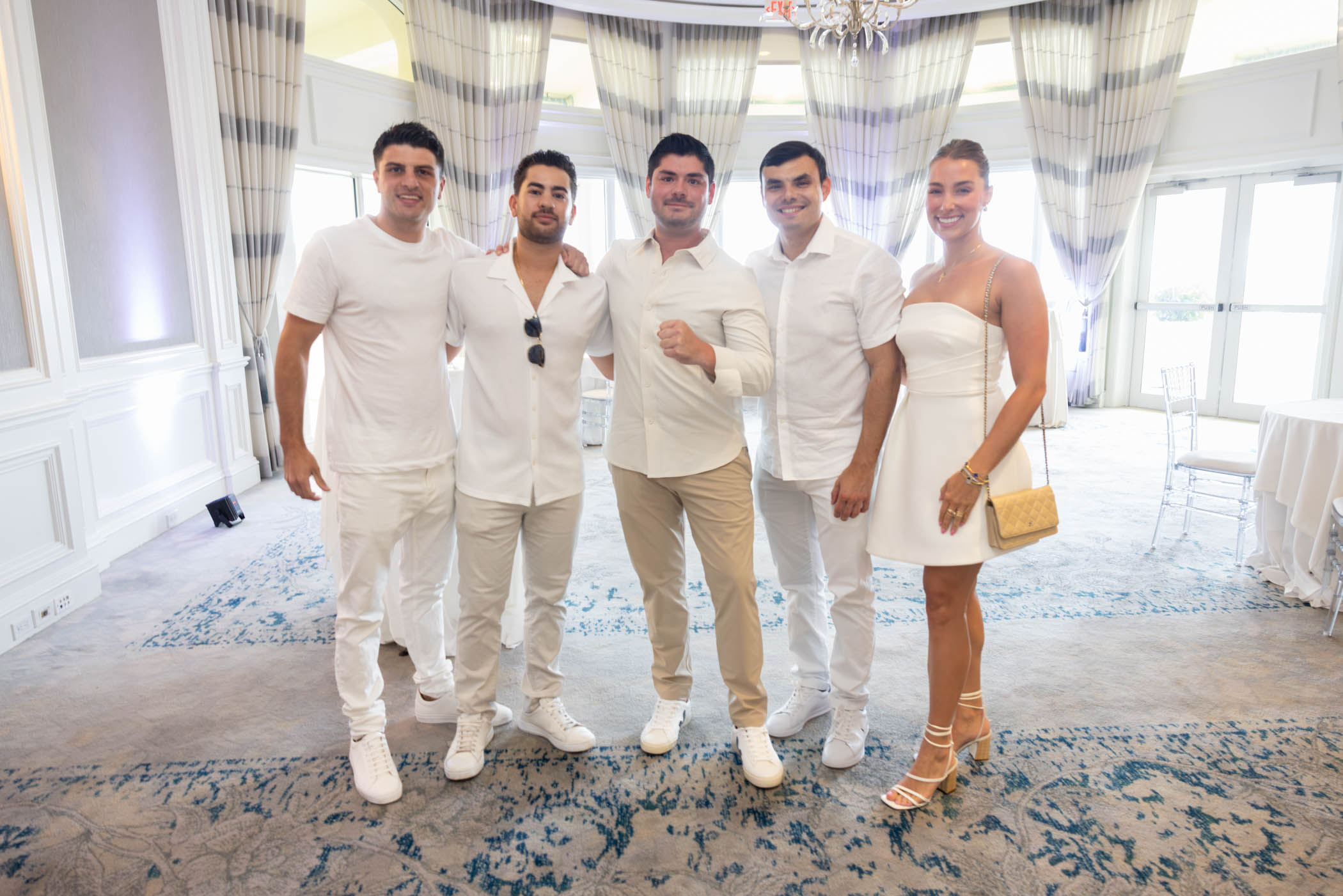 Five people pose together in a bright, elegant room with light curtains and blue carpet. Four men wear all white outfits, and one woman wears a white dress with strappy heels and holds a gold bag.