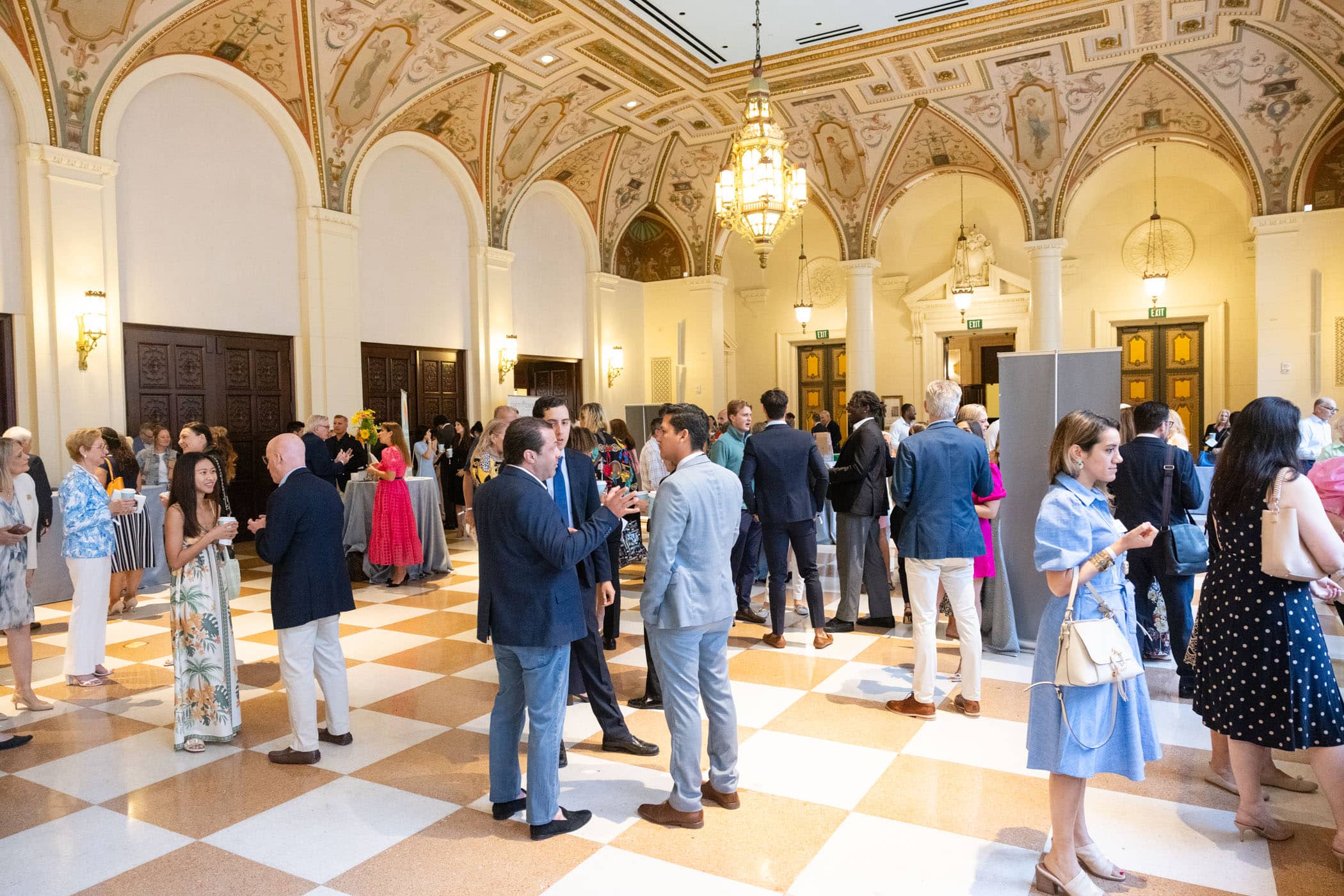 Guests in formal attire mingle and converse in an ornate hall with high ceilings, arched doorways, and elaborate frescoes. The room is bright and bustling with small groups gathered across the space.