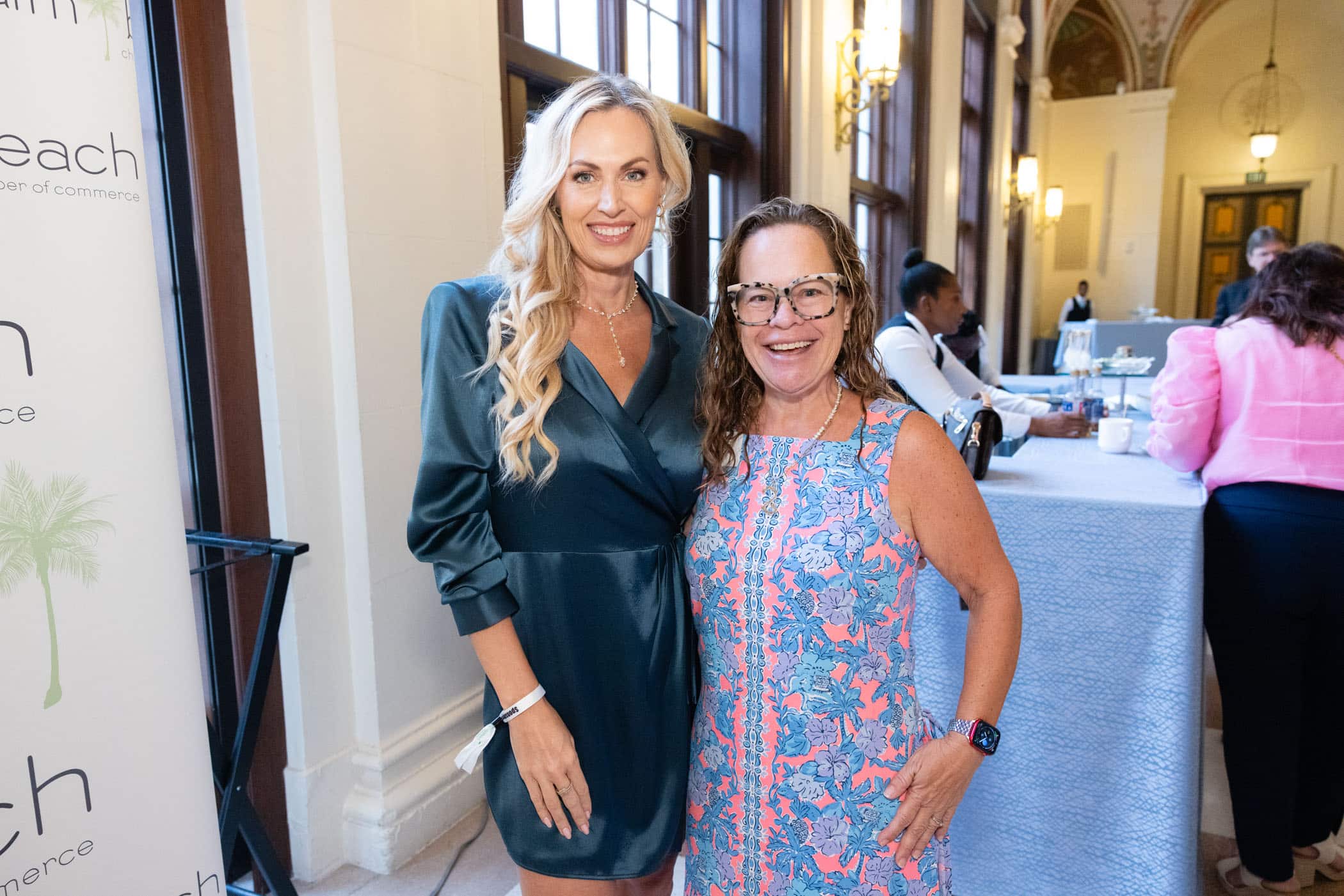 Two women stand smiling together indoors at an event. One wears a teal wrap dress and the other wears a colorful, sleeveless dress with glasses. People and tables are visible in the background.