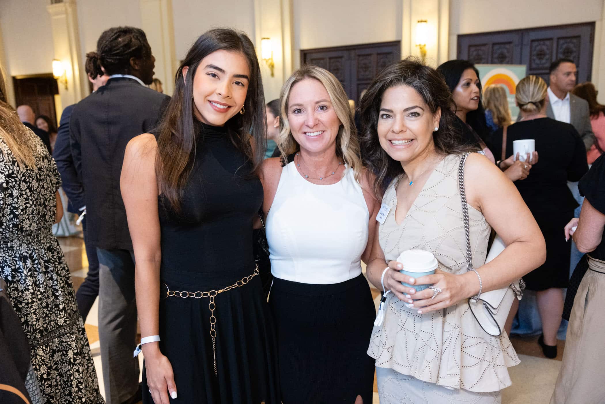 Three women stand close together, smiling at the camera during a crowded indoor event. One woman holds a cup, and other attendees are visible in the background, dressed in semi-formal attire.