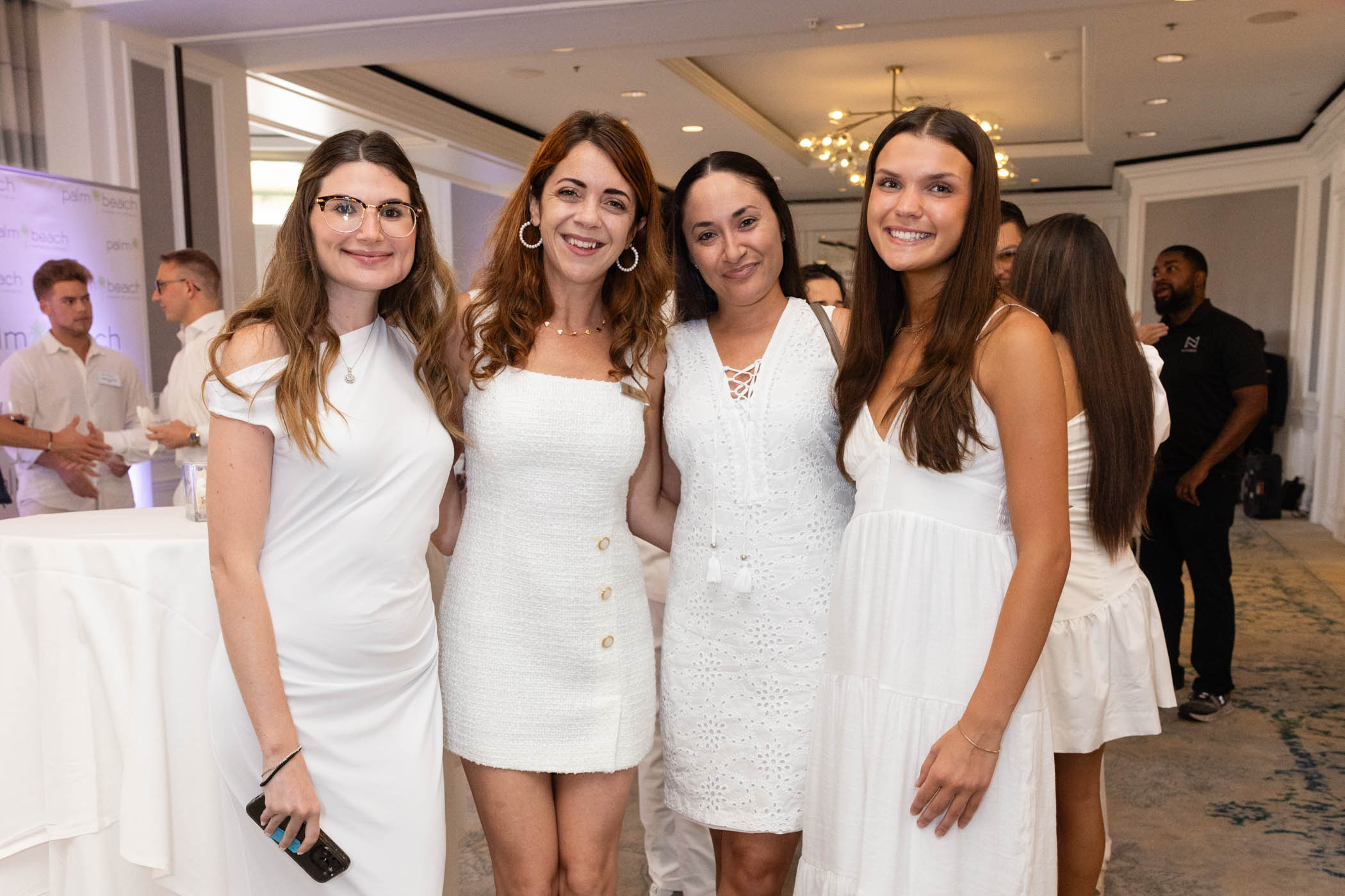 Four women wearing white dresses pose together and smile at a bright indoor event, with other people socializing in the background.