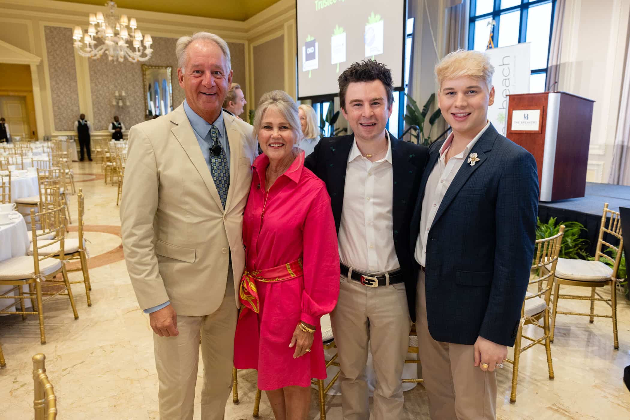 Four people stand together smiling at a formal event in a spacious, elegant room with gold chairs and chandeliers. Three men wear jackets and one woman wears a bright pink dress with a colorful belt.