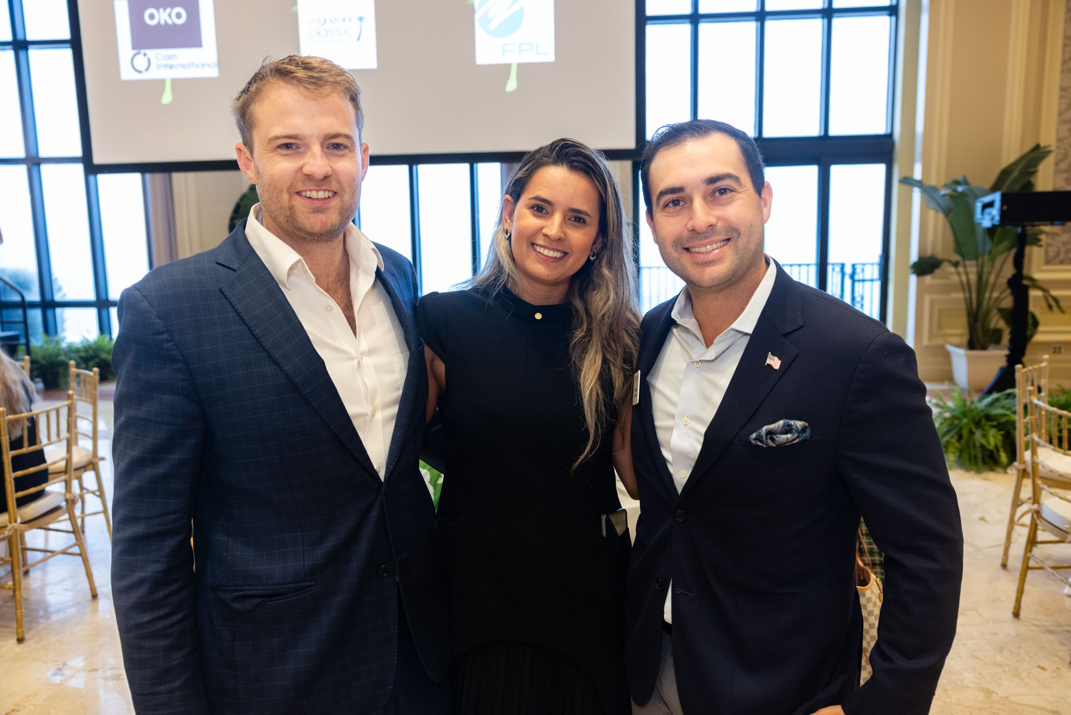 Three people—two men in suits and a woman in a black dress—stand smiling together indoors at an event, with large windows and a projection screen visible in the background.