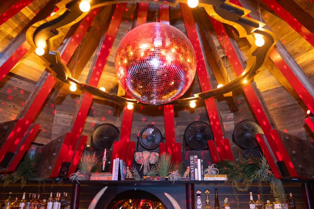 A shiny disco ball hangs from a wooden ceiling with red beams and warm string lights, reflecting spots of light across a bar decorated with books, plants, and fans.