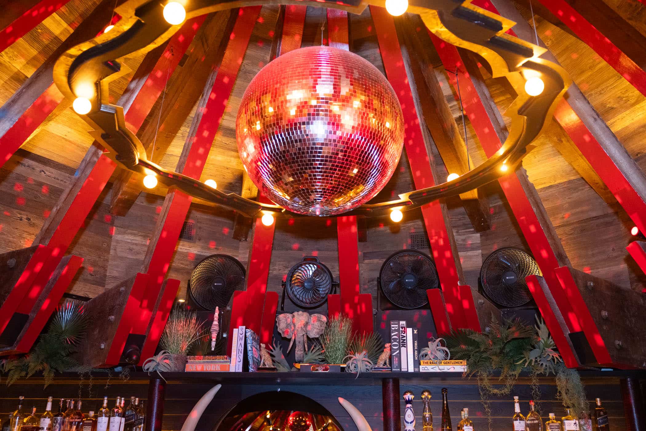 A shiny disco ball hangs from a wooden ceiling with red beams and warm string lights, reflecting spots of light across a bar decorated with books, plants, and fans.