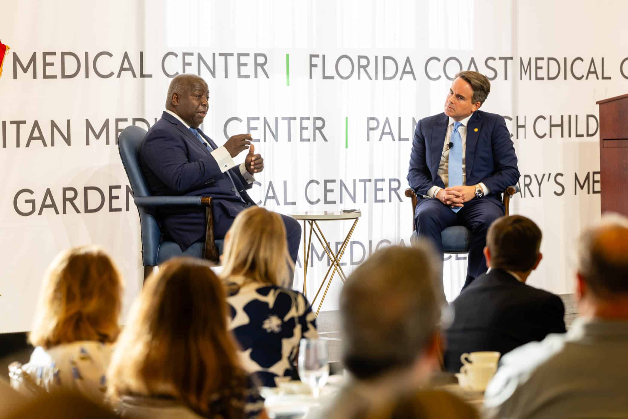 Two men in suits sit and talk on a stage in front of an audience, with a medical center banner in the background. One man gestures with his hands while speaking as the other listens attentively.