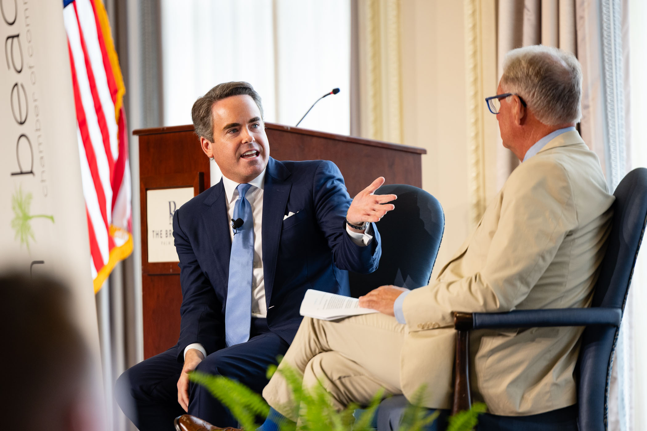 Two men in suits sit on stage having a lively conversation. One gestures with his hand while speaking. An American flag and a lectern are in the background, with audience members out of view.