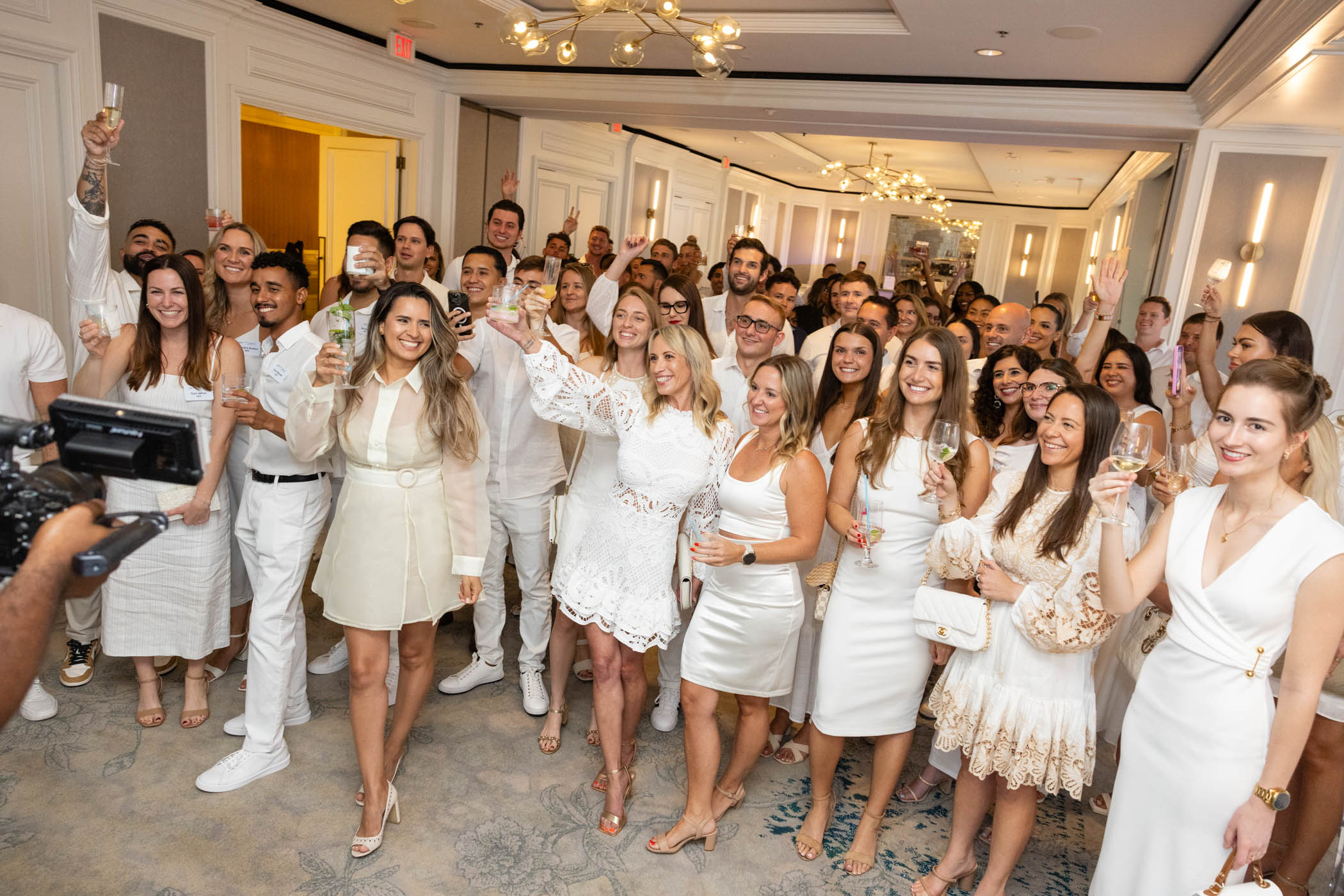 A large group of people dressed in white and cream attire smile and raise glasses in a toast at an indoor celebration in a well-lit, elegant room.