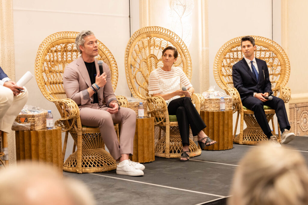 Three people sit on wicker chairs on a stage, engaged in a panel discussion. The man on the left speaks into a microphone, while the woman and man beside him listen attentively. Audience members are visible in the foreground.