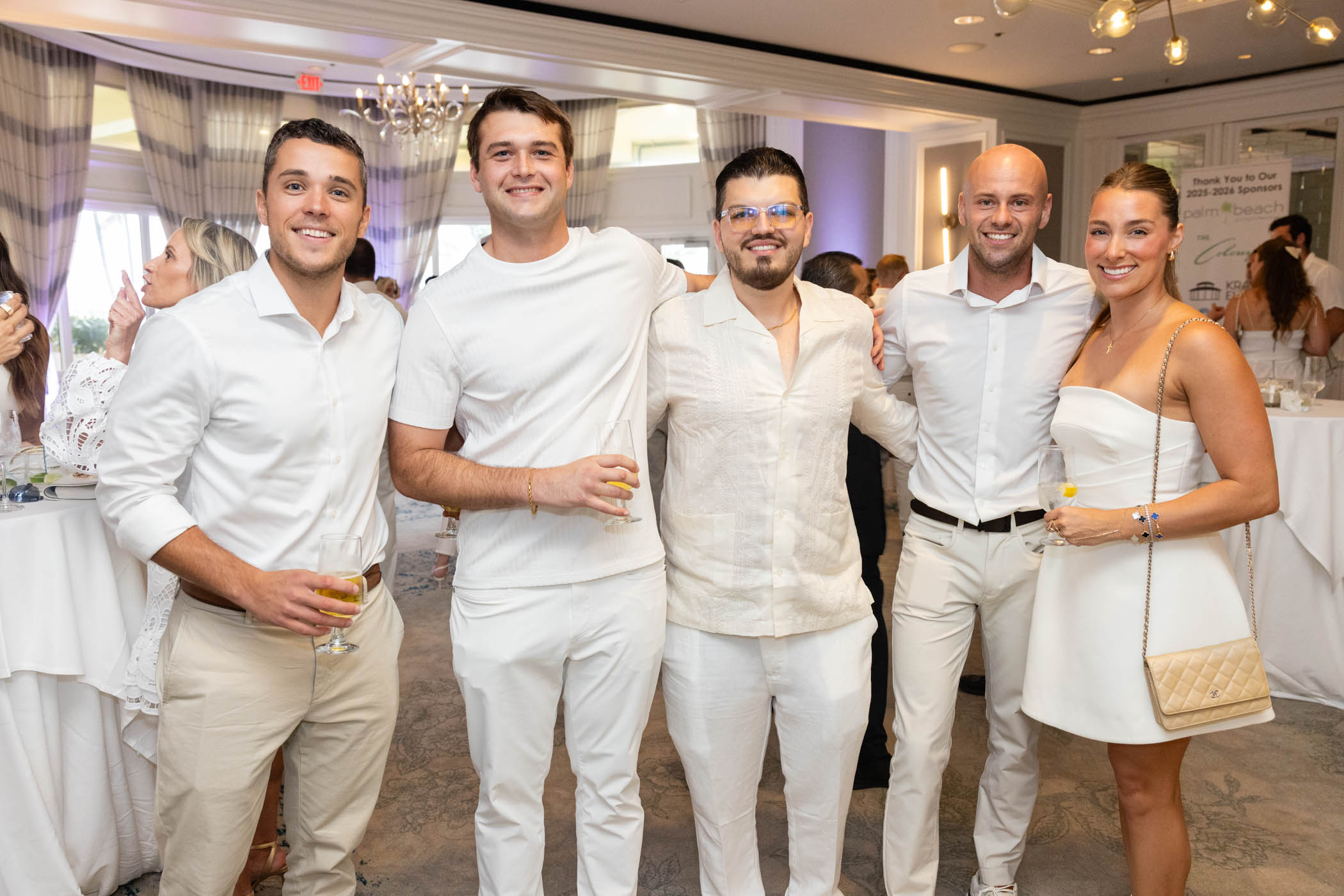 Five people dressed in white and beige outfits stand together smiling at an indoor event with drinks in hand. The setting is elegant, with chandeliers, draped curtains, and round tables in the background.