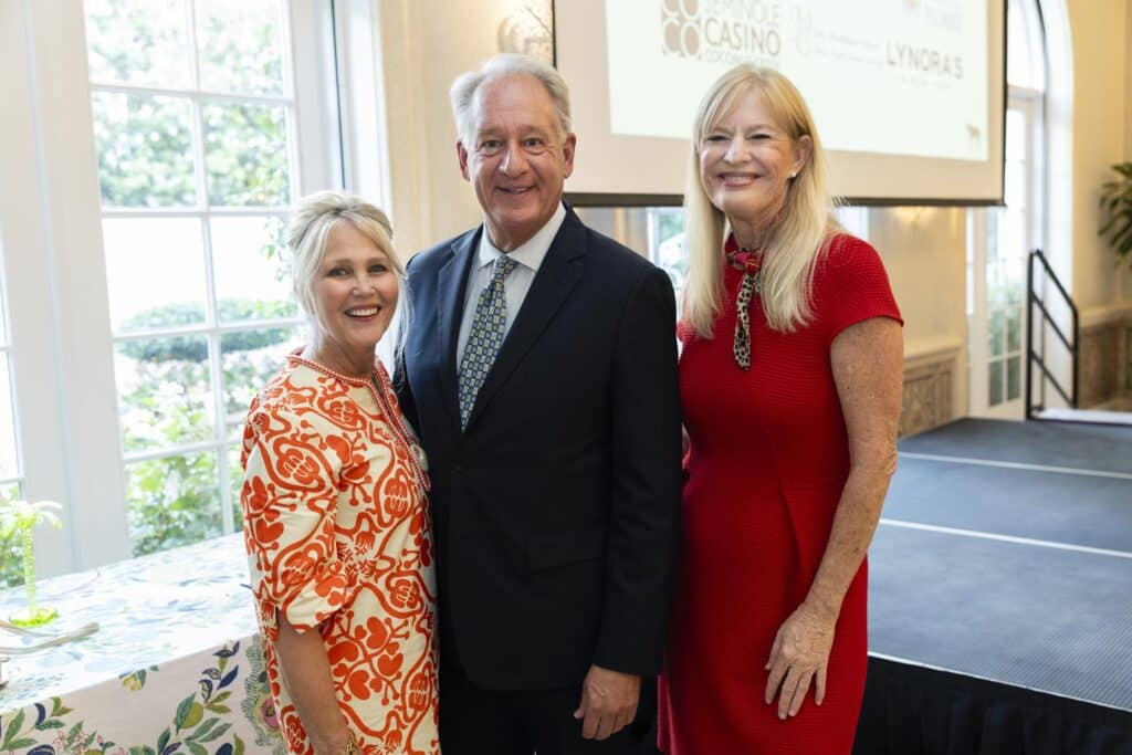 Three adults, two women and one man, stand smiling together indoors by a small round table. The women wear red dresses and the man is in a dark suit. A large window and a projection screen are visible in the background.