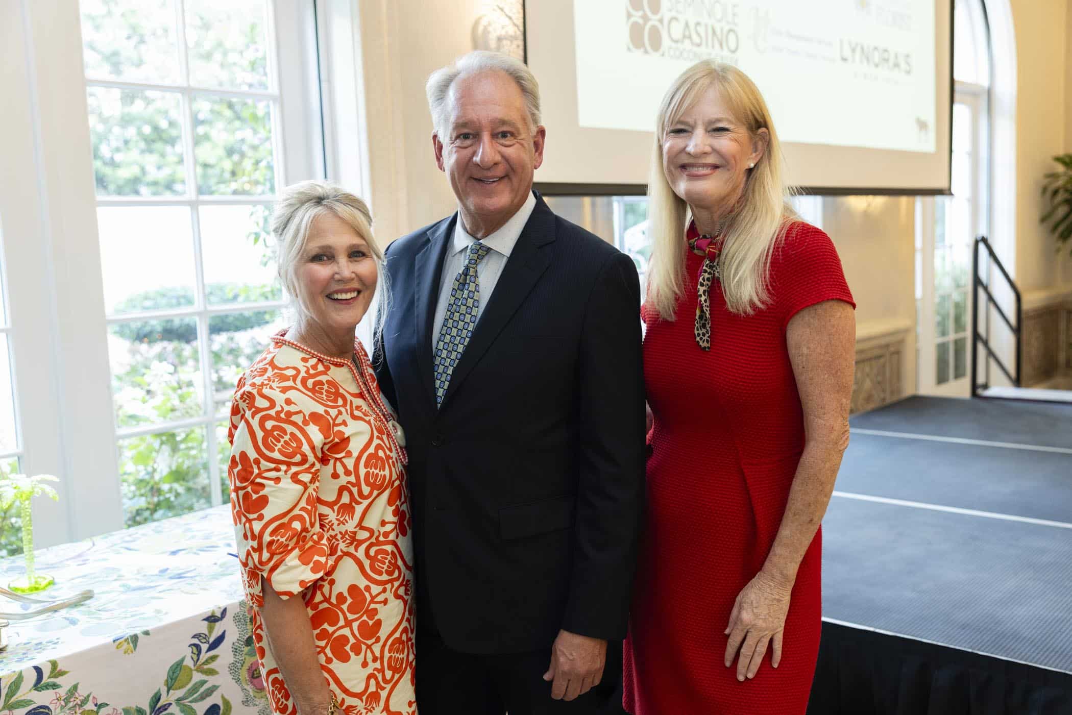 Three adults, two women and one man, stand smiling together indoors by a small round table. The women wear red dresses and the man is in a dark suit. A large window and a projection screen are visible in the background.