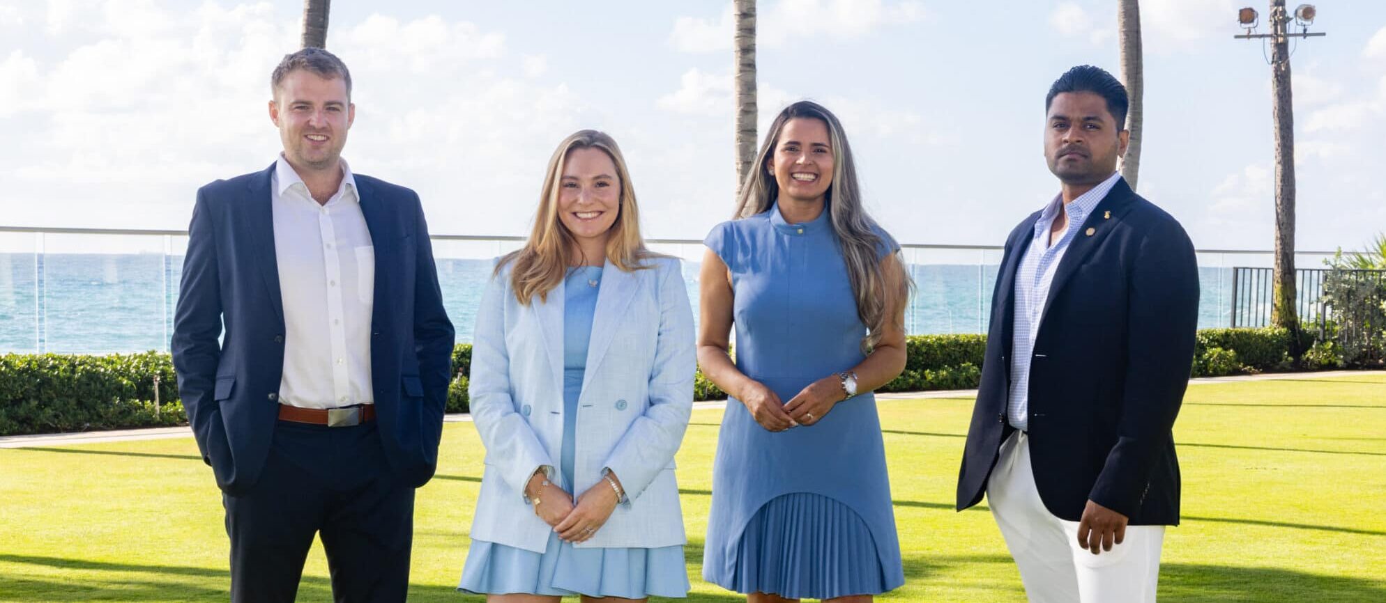 Four professionally dressed people stand on a lawn by the ocean, with palm trees and blue sky in the background. Two women wear light blue dresses, two men wear suits, and all are smiling at the camera.