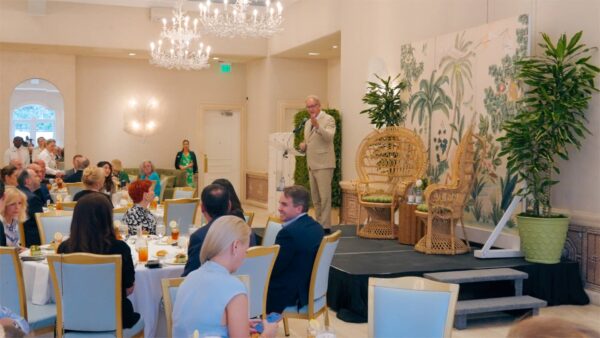 A man in a suit speaks on a stage with wicker chairs and tropical decor behind him, facing an audience seated at round tables in a brightly lit, elegant banquet room.