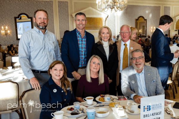 A group of seven people, dressed in business attire, pose and smile for a photo at a breakfast event. Breakfast foods and drinks are on the table, and a “Palm Beach Chamber of Commerce” logo is visible in the corner.