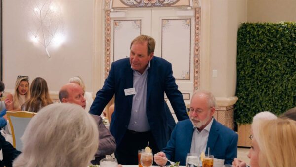 A man in a blue suit stands and speaks with seated guests at a formal dining event. People are engaged in conversation around tables set with drinks and salads, in a well-lit, elegant room with decorative walls.