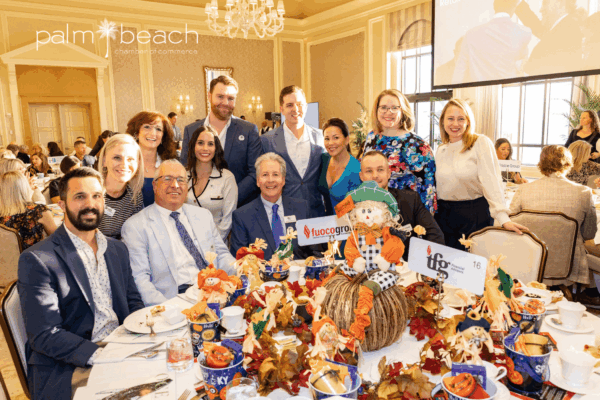 A group of people dressed in business attire smile around a decorated banquet table with autumn-themed centerpieces at a formal indoor event. The "Palm Beach Chamber of Commerce" logo is visible in the top left corner.