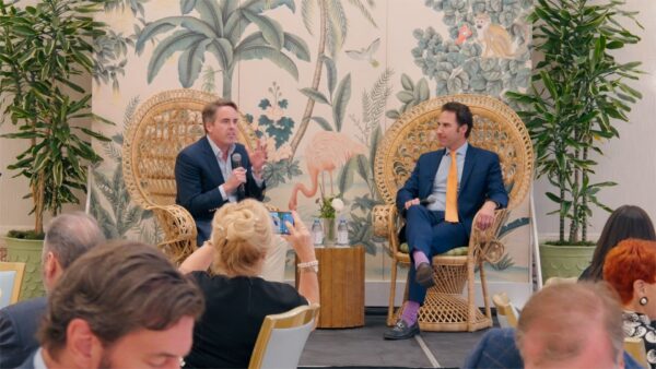 Two men in suits sit on wicker chairs with microphones, engaged in discussion on a stage decorated with tropical plants and a mural. Audience members watch and take photos in a well-lit, formal setting.