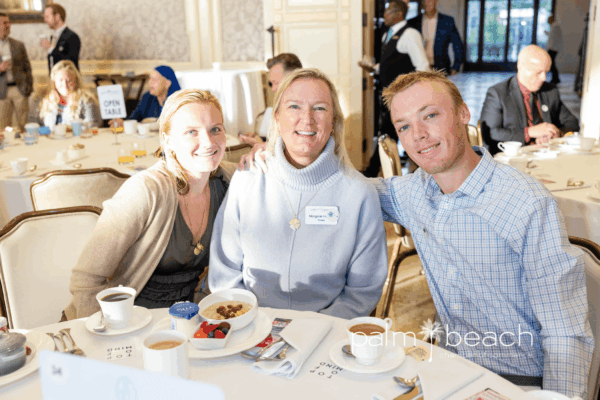 Three people sit closely together at a round table set for breakfast, smiling at the camera in a bright, elegant dining room with other guests and tables in the background. The "palm beach" logo is visible in the corner.
