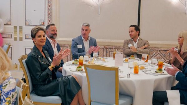 Six people in formal attire sit around a round table, clapping and smiling at a formal event. Salads, drinks, and plates are on the table in a brightly lit, elegant room.