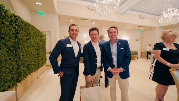 Three men in suits stand smiling together in a bright, elegant room with chandeliers and green wall decor. A woman in a black dress stands on the right, partially out of frame. The atmosphere appears formal and social.