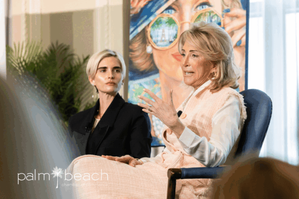 Two women are seated and engaged in conversation at an indoor event. One woman gestures while speaking. The background features a colorful portrait and the "Palm Beach Chamber of Commerce" logo is visible in the foreground.